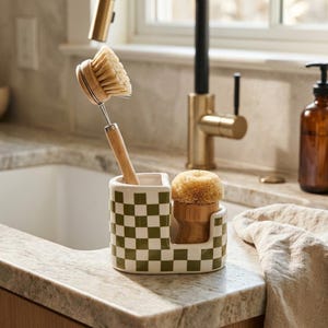 May include: A ceramic kitchen sink caddy featuring a green and white checkered pattern. The caddy holds two wooden dish brushes with light-colored natural bristles. One brush has a long handle, while the other is a compact, round palm brush.