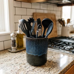 May include: A cylindrical ceramic utensil holder with a ribbed texture and a dark black base that transitions into a vibrant blue drip glaze at the top. The holder is filled with various black kitchen tools, including a slotted turner, whisk, and ladle.