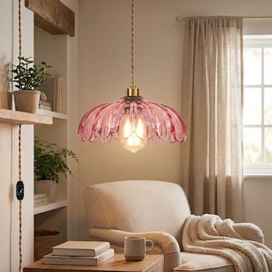 May include: A pink glass pendant light with a scalloped shade hangs from a brown cord. The light fixture has a brass-colored top and a vintage-style bulb. The light is in a cozy living room setting.