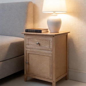 May include: A light-colored wooden nightstand with a drawer and a cabinet door. A white lamp with a lampshade sits on top, next to a closed book. The nightstand is next to a bed with a gray headboard.