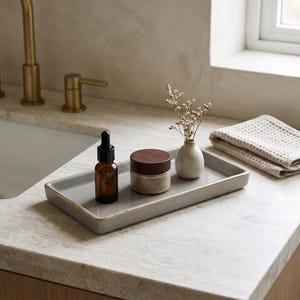 May include: A rectangular gray ceramic tray with a gold-toned rim holds a small amber glass dropper bottle, a round jar with a dark wooden lid, and a tiny speckled ceramic vase containing a small sprig of dried white flowers.