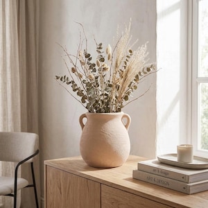 May include: A light beige, textured ceramic vase with two side handles, filled with a dried floral arrangement consisting of eucalyptus branches, tall grasses, and small white flowers, creating a neutral and organic decorative display.