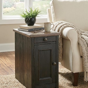 May include: A dark brown wooden side table with a drawer and cabinet door. A small potted plant, books, a mug, and eyeglasses sit on top. The table is next to a cream-colored armchair with a beige throw blanket.