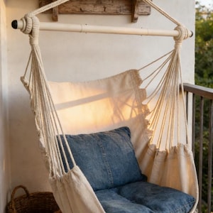 May include: A hanging chair made of off-white fabric and rope, with a denim blue cushion. The chair is suspended from a wooden bar and rope, and hangs on a balcony. A woven basket sits on the floor.