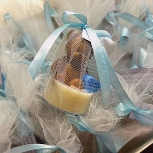 May include: A collection of baby shower favors. Each favor is a small, yellow container with a teddy bear and other small items inside, wrapped in white tulle and tied with a light blue ribbon.