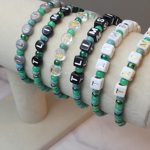 Puede incluir: Una colección de pulseras de cuentas de varios colores. Las pulseras presentan cuentas cúbicas con letras que deletrean palabras, intercaladas con cuentas redondas en verde, plata, negro y transparente. Las pulseras se exhiben sobre una superficie de tono neutro.