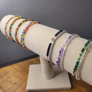 Op de afbeelding: Een display van kralenarmbanden in verschillende kleuren, waaronder goud, groen, oranje en paars. De armbanden zijn geregen op een zilveren draad en worden getoond op een witte cilindrische standaard. De achtergrond is neutraal grijs.