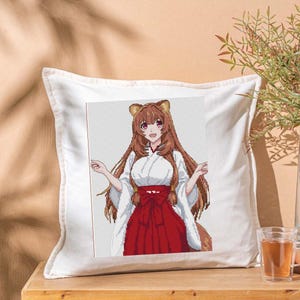 Puede incluir: Cojín cuadrado blanco con un diseño de punto de cruz de un personaje de anime con pelo largo castaño, que lleva un kimono blanco y una falda roja. El personaje tiene orejas de oso. El cojín está sobre una superficie de madera.