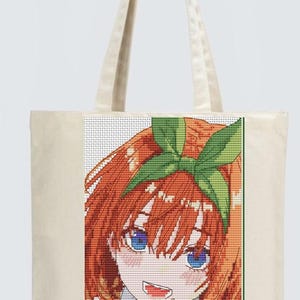 Puede incluir: Un bolso tote beige con un diseño de punto de cruz colorido de un personaje de anime con cabello naranja, ojos azules y un lazo verde. El personaje sonríe y hace un signo de paz. El bolso tiene dos asas beige.