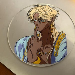 Peut inclure: Une peinture sur verre circulaire représentant un personnage de style anime aux cheveux blonds, à la peau foncée et aux yeux bleus. Le personnage porte un vêtement bleu et doré à motifs, une boucle d'oreille ankh dorée et un collier doré.