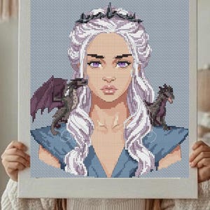 May include: Cross-stitch portrait of a woman with long, white hair, a crown, and two small dragons. The artwork is framed in a light-colored wooden frame. The woman wears a blue dress and a gold necklace.