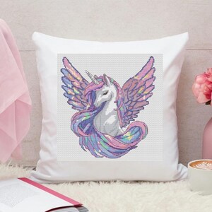 May include: White square pillow with a cross-stitch design of a unicorn with wings. The unicorn has a white body, a pink and purple mane and tail, and iridescent wings. The pillow is on a white surface.