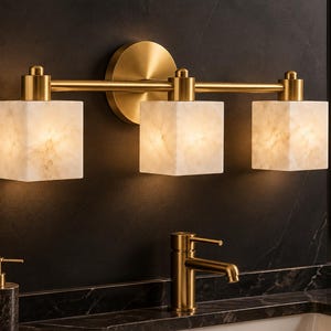 May include: A three-light vanity wall fixture featuring a brushed brass horizontal bar and circular wall mount. It includes three square, translucent white stone shades with a marbled texture that emit a warm, soft glow when illuminated.