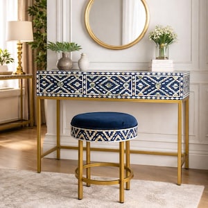 Puede incluir: Una mesa de tocador azul marino con patas doradas y un taburete a juego. La mesa tiene tres cajones con un patrón geométrico. Un espejo con marco dorado cuelga encima y un jarrón con flores está encima.