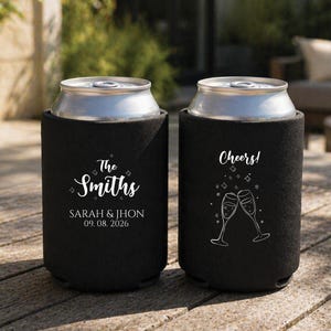 May include: Two black foam can coolers featuring white printed designs. One cooler displays the text The Smiths, Sarah & Jhon, and the date 09.08.2026. The other cooler features the word Cheers! and a graphic of two clinking champagne flutes.