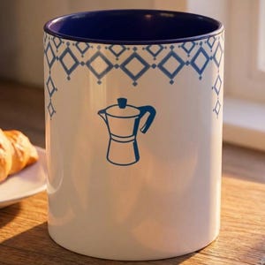 Könnte beinhalten: Eine weiße Keramik-Tasse mit schwarzem Henkel und dem Text "But First, Coffee" in fetter, schwarzer Schrift. Die Tasse steht vor einem verschwommenen Hintergrund, der eine gemütliche Umgebung suggeriert.