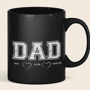 Op de afbeelding: Zwarte keramische mok met het woord "DAD" in witte, verweerde blokletters. Onder het woord "DAD" staan drie namen: Max, Kevin en Marlene, elk met een hartsymbool.
