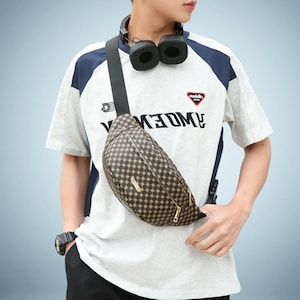 May include: A brown and tan checkered fanny pack with a gold zip. The bag is worn across the body. The person is wearing a white t-shirt with navy blue accents and the word "KEDM" printed on it.