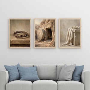 May include: Three framed art prints depicting religious themes. One shows a crown of thorns, another a tomb with a stone, and the third a draped cloth. The prints are in neutral tones and are displayed above a gray sofa with blue pillows.