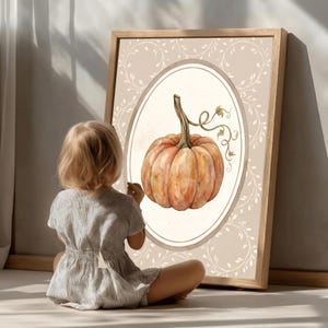 Puede incluir: Una l&aacute;mina de arte enmarcada que presenta una ilustraci&oacute;n en acuarela de una calabaza naranja con tallo verde y zarcillos, centrada dentro de un borde ovalado sobre un fondo beige claro decorado con delicados patrones de enredaderas blancas.