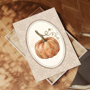 Puede incluir: Una impresi&oacute;n rectangular que presenta una ilustraci&oacute;n en acuarela de una calabaza naranja con un tallo verde y zarcillos. La calabaza est&aacute; centrada dentro de un marco ovalado sobre un fondo beige claro decorado con un patr&oacute;n de hojas blancas.