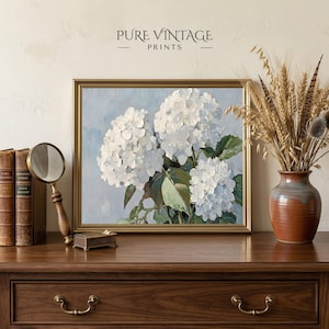 Puede incluir: Una impresi&oacute;n floral enmarcada con hortensias blancas sobre un fondo azul claro. La obra de arte est&aacute; en un marco dorado y se exhibe sobre una superficie de madera con otros elementos decorativos, incluyendo una lupa y un jarr&oacute;n con plantas secas. El texto "PURE VINTAGE PRINTS" est&aacute; en la parte superior.