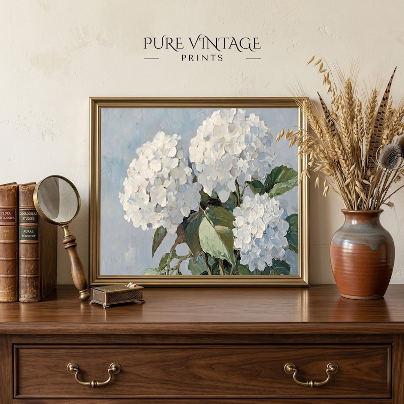 Puede incluir: Una impresi&oacute;n floral enmarcada con hortensias blancas sobre un fondo azul claro. La obra de arte est&aacute; en un marco dorado y se exhibe sobre una superficie de madera con otros elementos decorativos, incluyendo una lupa y un jarr&oacute;n con plantas secas. El texto "PURE VINTAGE PRINTS" est&aacute; en la parte superior.