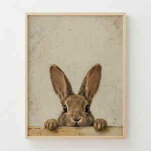May include: A framed print featuring a brown rabbit with long ears, peeking over a wooden ledge. The rabbit's fur is detailed, and the background is a textured, neutral color. The frame is a light wood tone.