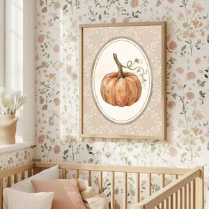 Puede incluir: Una impresi&oacute;n de arte mural enmarcada que presenta una ilustraci&oacute;n en acuarela de una calabaza naranja centrada dentro de un borde ovalado. El fondo de la impresi&oacute;n es de color beige claro decorado con delicados patrones de enredaderas blancas.