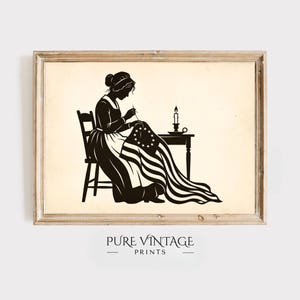 May include: A framed print featuring a silhouette of a woman seated, sewing a flag with stars and stripes. A candle sits on a table beside her. The print has a vintage aesthetic, with the text "PURE VINTAGE PRINTS" at the bottom.