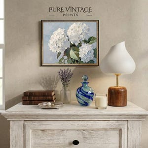 Puede incluir: Una impresi&oacute;n floral enmarcada con hortensias blancas cuelga sobre una c&oacute;moda blanca. Una l&aacute;mpara blanca con base de madera, una botella de vidrio azul y verde y una peque&ntilde;a vela est&aacute;n sobre la c&oacute;moda. El texto "PURE VINTAGE PRINTS" est&aacute; en la parte superior.