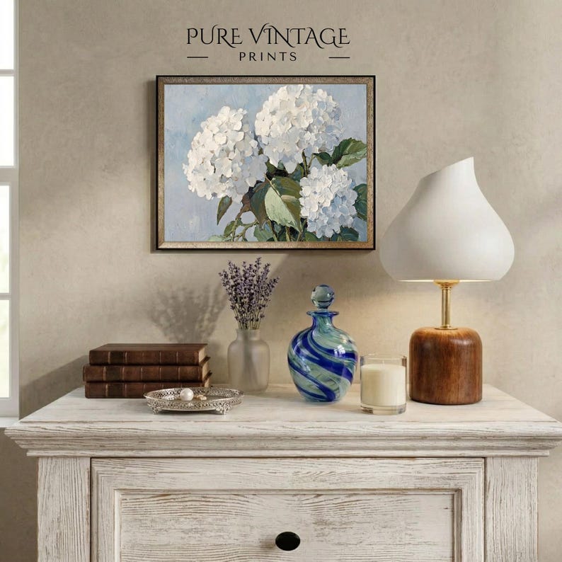 Puede incluir: Una impresi&oacute;n floral enmarcada con hortensias blancas cuelga sobre una c&oacute;moda blanca. Una l&aacute;mpara blanca con base de madera, una botella de vidrio azul y verde y una peque&ntilde;a vela est&aacute;n sobre la c&oacute;moda. El texto "PURE VINTAGE PRINTS" est&aacute; en la parte superior.