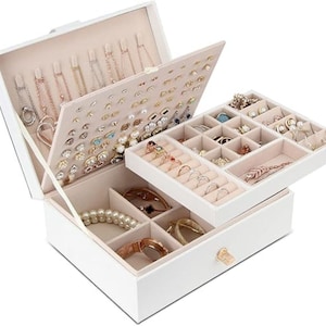 May include: A white jewellery box with multiple compartments and drawers. The box is open, revealing various necklaces, rings, earrings, bracelets, and other jewellery pieces. The interior is lined with a soft, light-coloured fabric.
