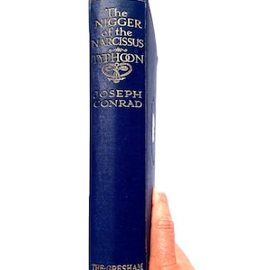 Può includere: Un libro vintage con copertina rigida intitolato "The Nigger of the Narcissus" di Joseph Conrad. Il libro ha una copertina blu scuro con scritte dorate e la parola "Typhoon" è visibile. Pubblicato da The Gresham Publishing Co. LTD.