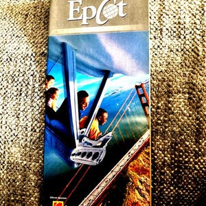 Può includere: Un GuideMap di Epcot vintage con il titolo in bianco e il logo Epcot. La copertina presenta un'immagine colorata di persone su un'attrazione con un ponte sullo sfondo. La guida è uno sponsor ufficiale di Eastman Kodak Company.