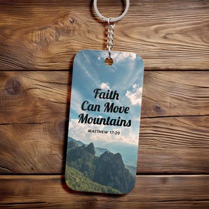 Puede incluir: Un llavero rectangular con un paisaje de montaña y un fondo de cielo. El texto "Faith Can Move Mountains" y el versículo bíblico Mateo 17:20 están impresos en la parte delantera. El llavero está sujeto a un llavero plateado.