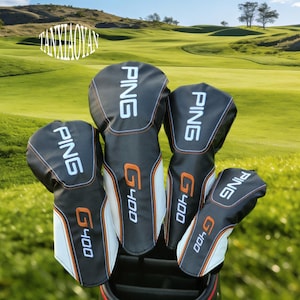 Puede incluir: Un juego de fundas para palos de golf en blanco y negro con la palabra "PING" en blanco y "G400" en naranja. Las fundas están dispuestas en una bolsa de golf, con un campo de golf verde de fondo.
