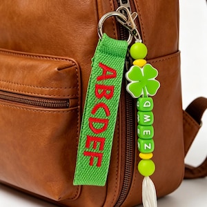 May include: A brown leather backpack featuring a green fabric keychain with the red embroidered letters ABCDEF and a beaded charm with a green four-leaf clover, yellow beads, green lettered cubes spelling ZEMQ, and a white tassel attached to the zip.