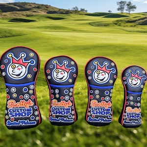 May include: Four black golf club head covers with a smiling jester face design, red and blue accents, and the text "Scotty's Custom Shop" and "Limited Release". The covers are on a green golf course.
