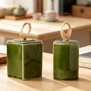 May include: Two glossy olive green ceramic canisters with lids, each featuring a natural jute twine loop handle on top. One canister is square-shaped, while the other is cylindrical. The text "Customizable gifts" is visible in red.