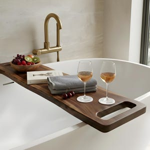 Puede incluir: Una bandeja de bañera de madera oscura con un cuenco de fruta, un libro, una toalla gris y dos copas de vino. La bandeja se extiende sobre una bañera blanca, con un grifo dorado en el fondo.