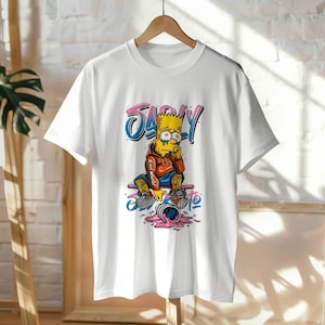 Puede incluir: Camiseta blanca con una ilustración de dibujos animados de Bart Simpson. Bart se muestra con una expresión triste, con una sudadera con capucha roja y sentado en una patineta con la palabra "SAD" encima en letras rosas y azules. La patineta tiene un diseño de helado rosa derretido.