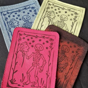 Op de afbeelding: Vier stoffen patches met unieke illustraties. De patches zijn in tinten blauw, geel, roze en bruin. Elke patch heeft een zwart-wit ontwerp van een skelet en figuren, omringd door bloemmotieven. De randen zijn gezoomd.