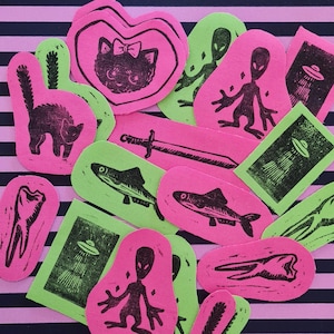 Op de afbeelding: Een verzameling neonroze en groene gestanste stickers met zwarte linoleumsnede ontwerpen. De stickers tonen verschillende afbeeldingen, waaronder een kat, aliens, vissen, een zwaard, een tand en een UFO.