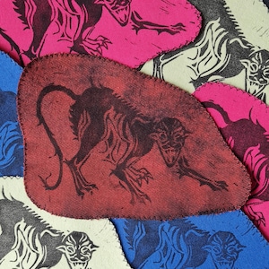 May include: A selection of fabric patches in shades of pink, red, blue, and cream. Each patch displays a black illustration of a snarling beast. The patches are irregularly shaped with a stitched edge, creating a textured appearance.