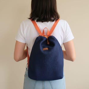 Op de afbeelding: Een marineblauwe rugzak met oranje bandjes wordt over de schouders gedragen. De rugzak heeft een geweven textuur en een klein bruin leren label. De persoon draagt een wit T-shirt en een blauwe spijkerbroek.