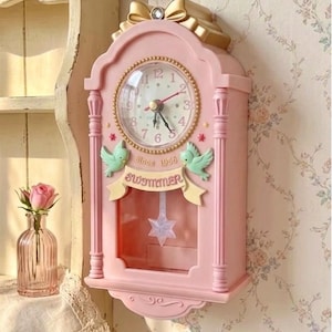 May include: A pink wall clock featuring a gold bow at the top, a circular clock face with numbers 1 through 12, two light green birds, and a banner with the text "Since 1986 SWIMMER". A star-shaped pendulum hangs behind a clear glass panel.