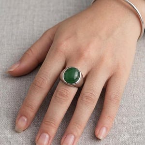 Peut inclure: Une bague argentée ornée d'une pierre ovale lisse de couleur vert foncé, montée sur un anneau en métal poli. La bague est portée à l'annulaire et un fin bracelet jonc en métal argenté assorti est visible au niveau du poignet.