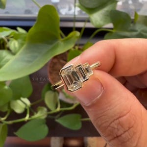 Può includere: Un anello d'oro con tre pietre preziose taglio smeraldo. L'anello ha una fascia sottile e un'incastonatura che mette in risalto la chiarezza delle pietre. L'anello è tenuto in mano, con il nome del marchio "SHYAM LUXE" visibile sullo sfondo.