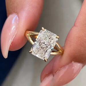 Op de afbeelding: Een gouden ring met een rechthoekig geslepen diamant. De diamant schittert helder. De ring heeft een split-schacht ontwerp. De band is gemaakt van goud, wat bijdraagt aan de elegantie van de ring.